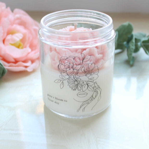 A scented candle from Grace and Bloom with a beautiful peach colored wax flower nestled inside the jar. A hand-drawn line drawing of a lady with most of her face covered in various flowers graces the front of the jar.