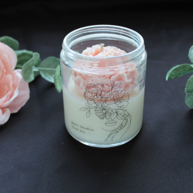 A scented candle from Grace and Bloom with a beautiful peach colored wax flower nestled inside the jar. A hand-drawn line drawing of a lady with most of her face covered in various flowers graces the front of the jar.