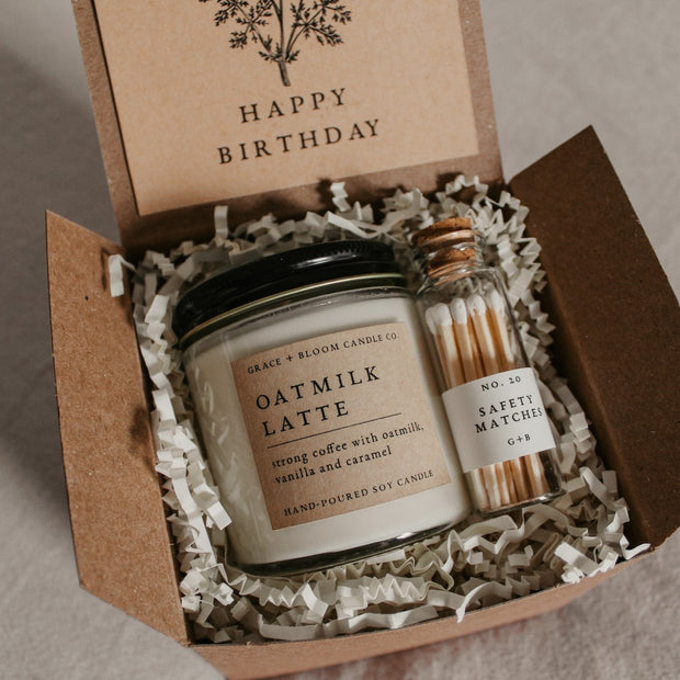 A cozy gift box from Grace and Bloom, perfect for bookish friends, has a candle in your choice of literary scents, a glass bottle of matches, and has a removable kraft-colored card with a floral design and your choice of messages on it.